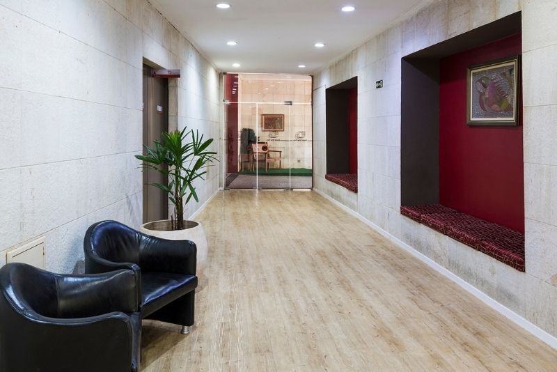 Corredor moderno com cadeiras pretas, paredes brancas, nichos vermelhos e um vaso de planta ao fundo.