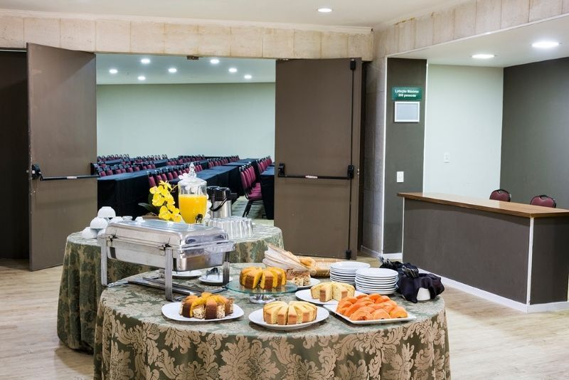 Mesa de buffet com doces, frutas e bebidas servidas em uma sala de conferências de hotel.