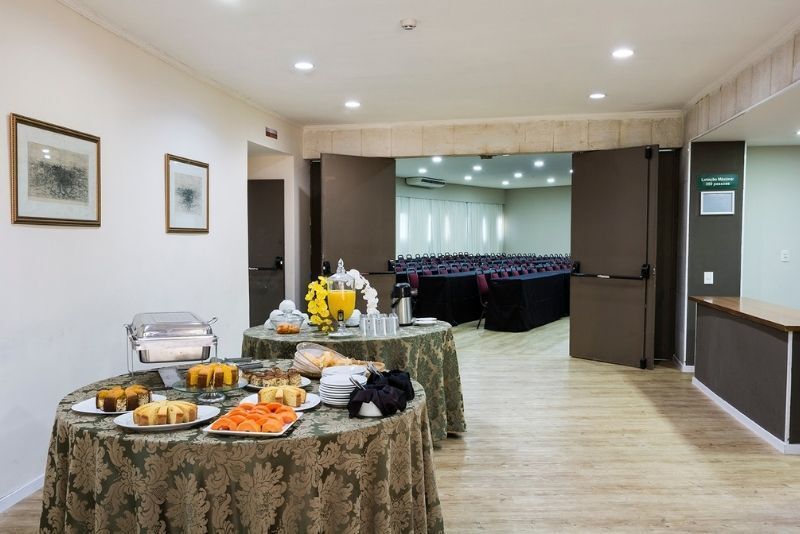 Buffet servido em uma sala de conferências iluminada, com mesas cobertas e portas duplas abertas.
