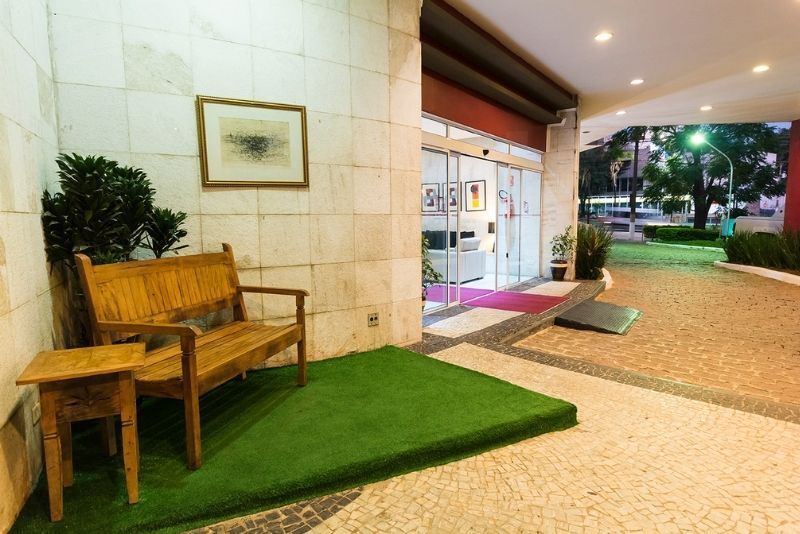 Banco de madeira ao lado de uma parede de azulejos sob uma entrada coberta, com grama verde e uma passarela que leva ao exterior.