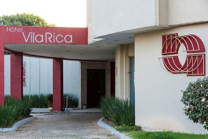 Entrada do Hotel Vila Rica com placa e logotipo vermelhos em um edifício branco.