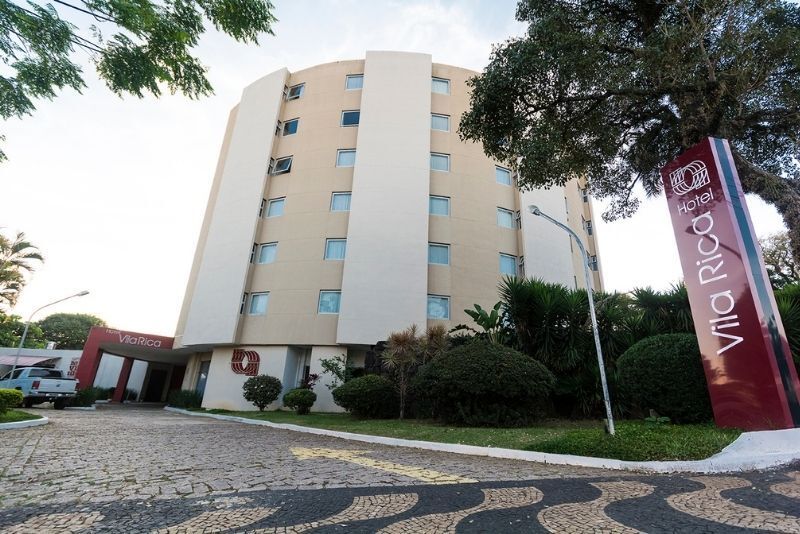 Prédio moderno de hotel em tons bege com uma placa vermelha, visto de uma entrada pavimentada rodeada por árvores.