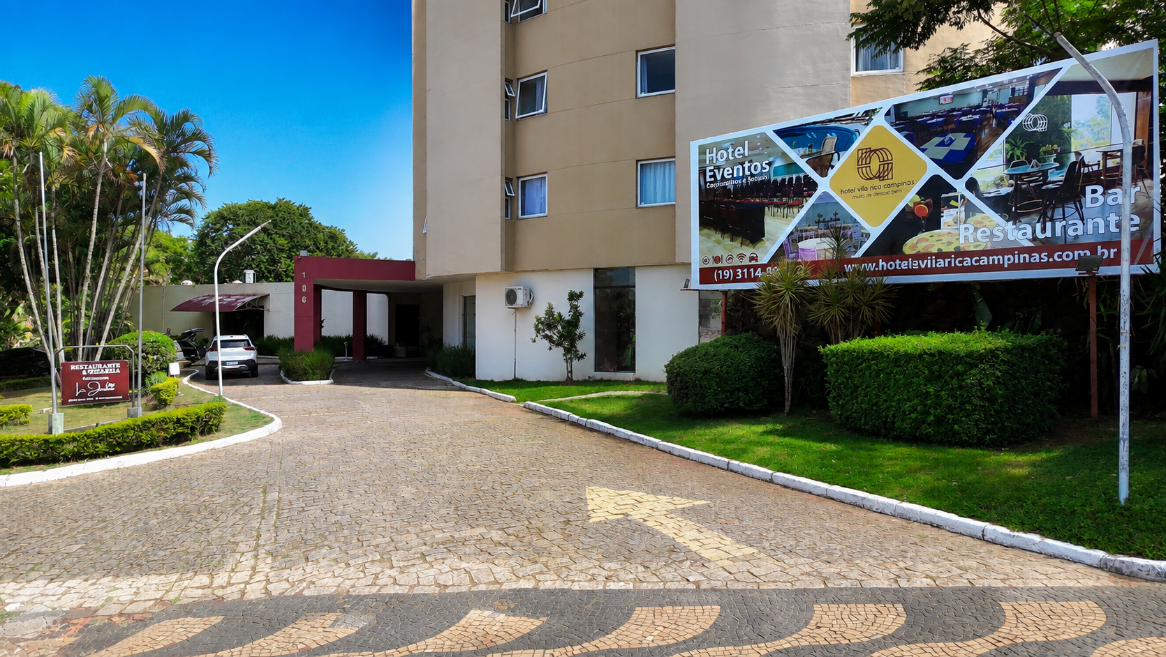 Entrada de hotel moderno ao entardecer, com fachada alta e curva, saguão iluminado, palmeiras e carro estacionado.