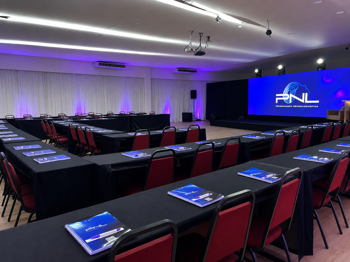 Amplo salão de conferências com fileiras de mesas voltadas para um palco iluminado e um telão.