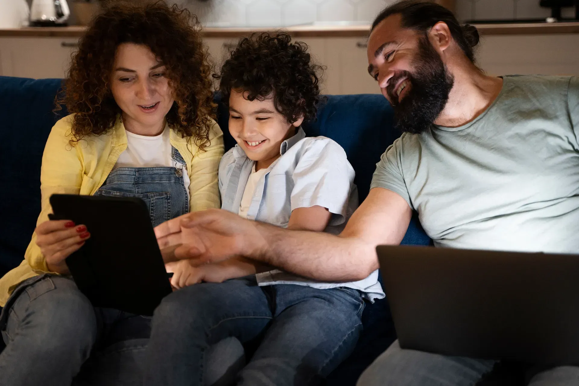 Três pessoas sentadas em um sofá, usando tablets e laptops, sorrindo juntas em uma sala de estar aconchegante.