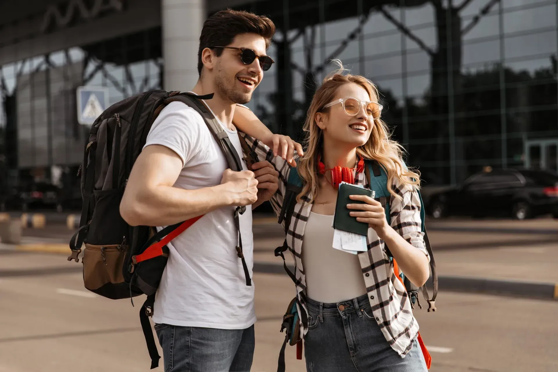 Dois viajantes sorridentes com mochilas, segurando garrafas de água e mapas, estão em frente a um prédio de vidro.