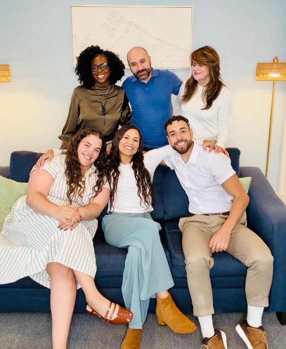 Six people sitting and standing in a bright living room, smiling for a group photo.