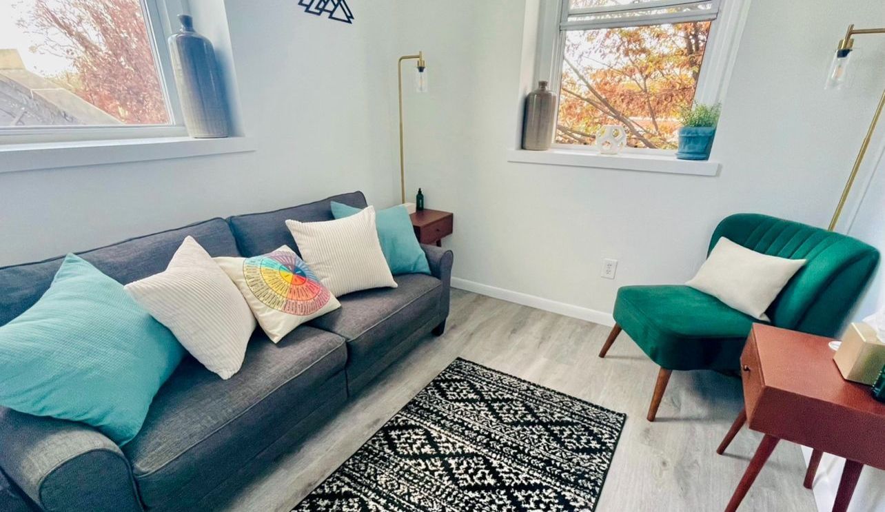 Living room with gray sofa, teal chair, rug, and small side tables. Window with fall foliage.