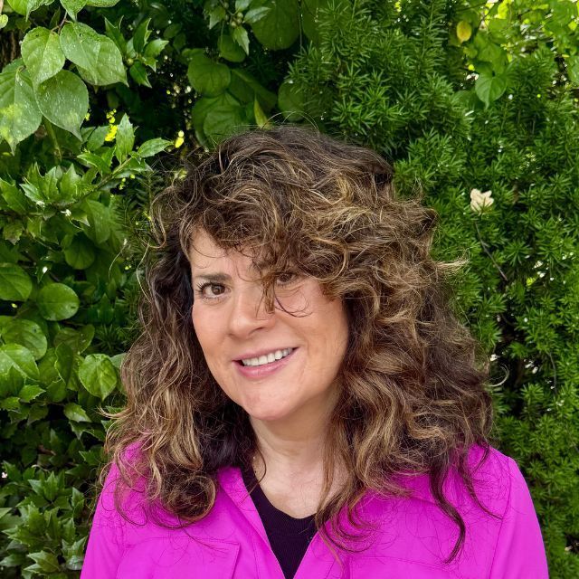 Woman with dark wavy hair smiling, green foliage background.