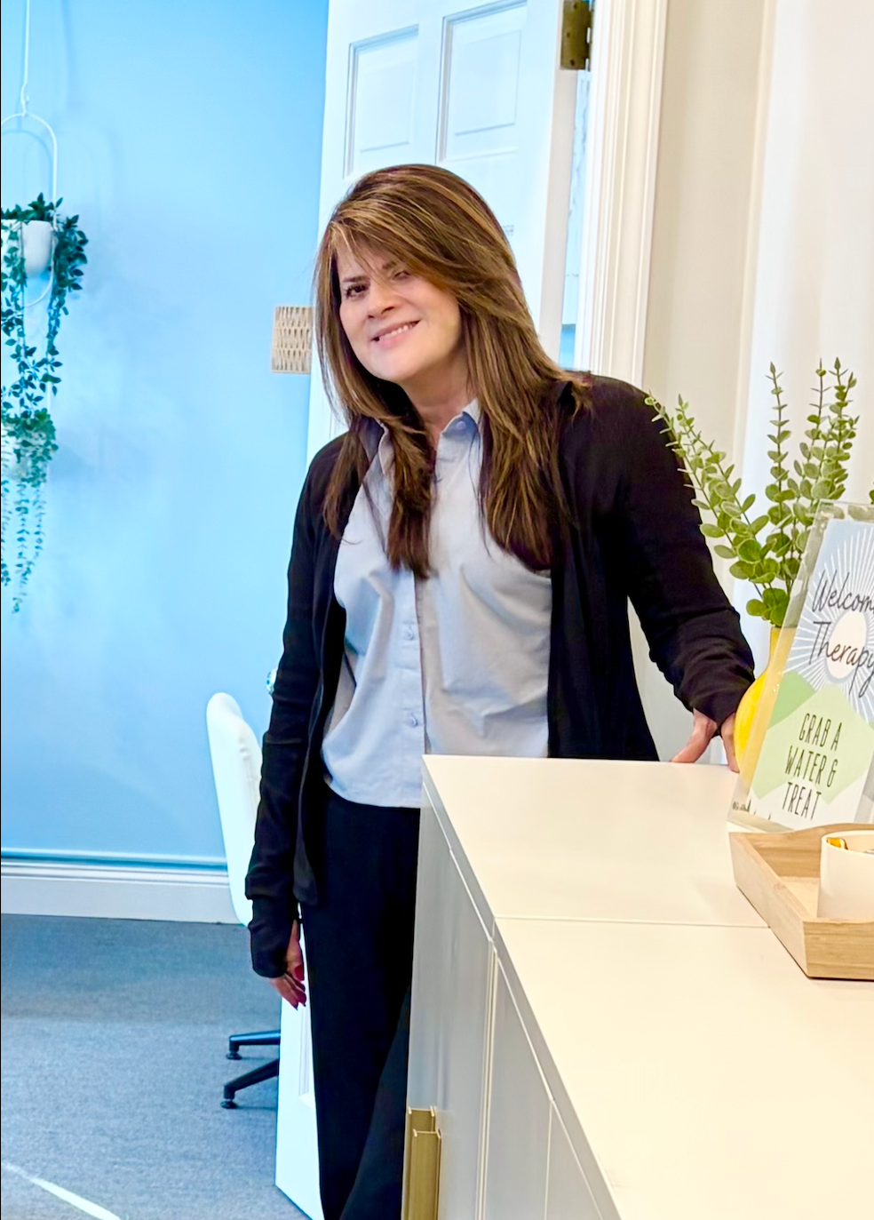 Woman standing by a white counter in a bright office, smiling and leaning slightly forward.