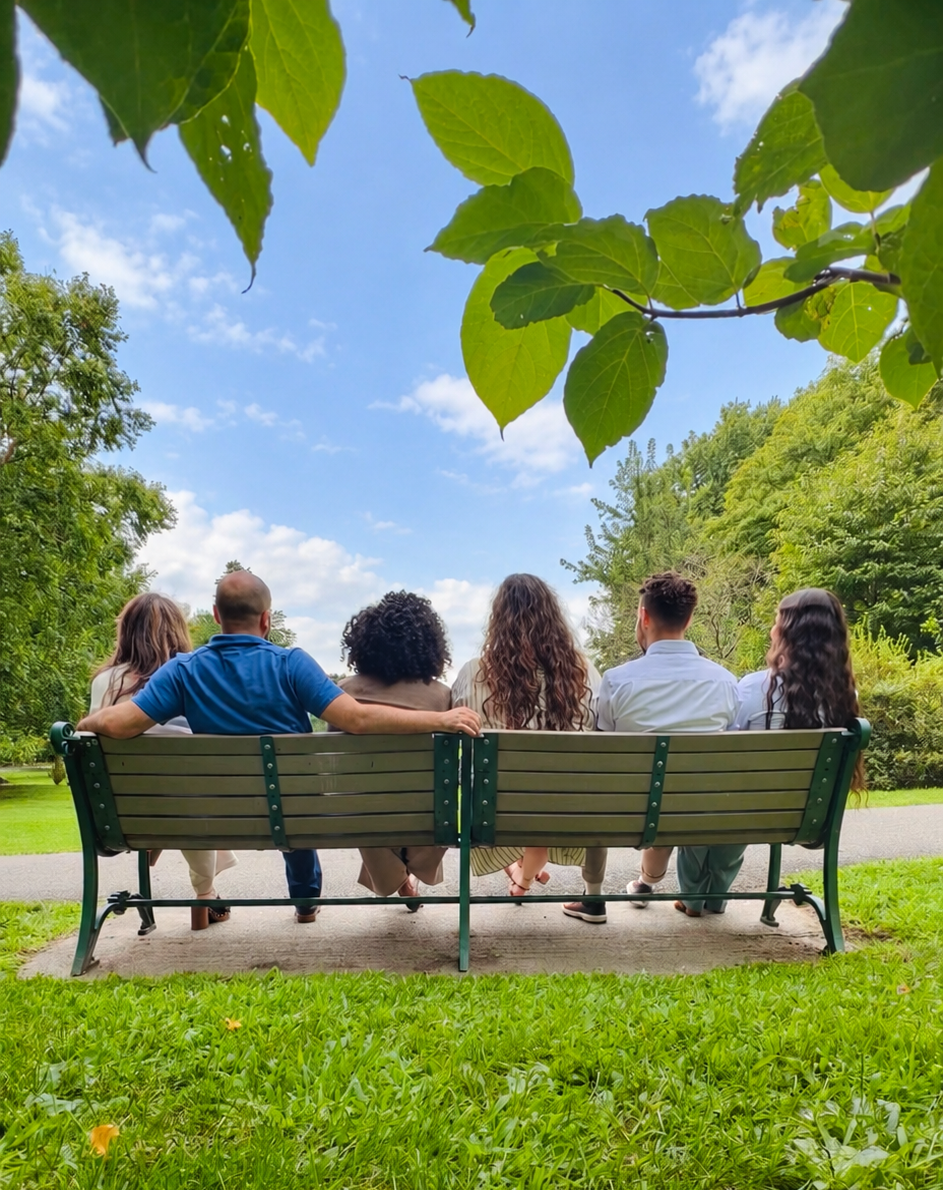 Five people sit on a park bench under green leaves, facing a sunny blue sky and grassy lawn.