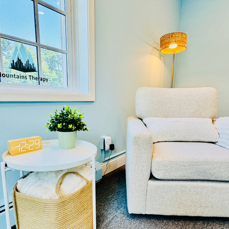 Bright therapy room with white armchair, side table, lamp, and a potted plant by a window