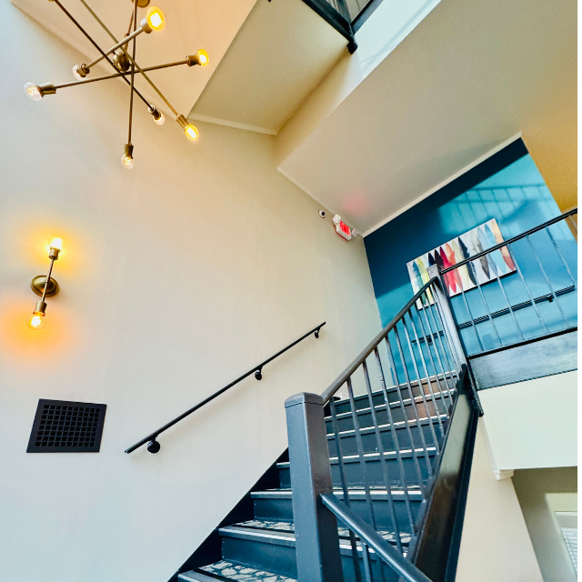 Staircase with black railing, gray walls, and modern lighting fixture. Abstract artwork and blue accent wall.