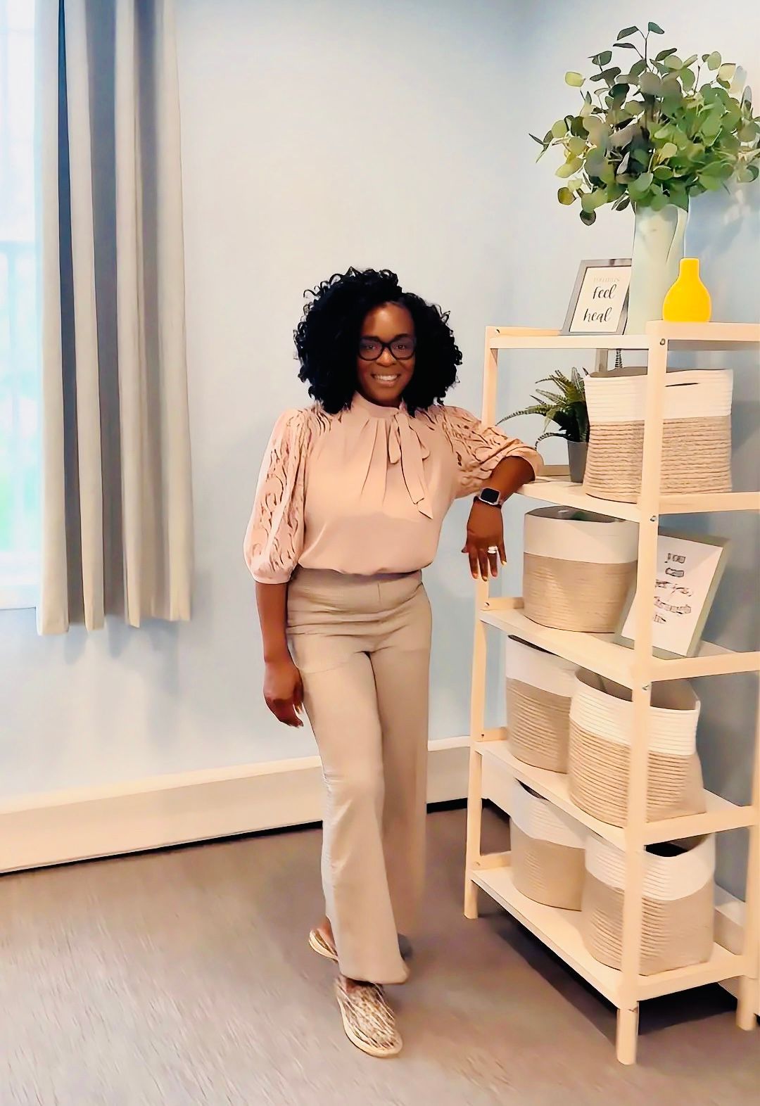 Woman standing beside a wicker shelf in a bright room, wearing beige clothes and leopard-print shoes.