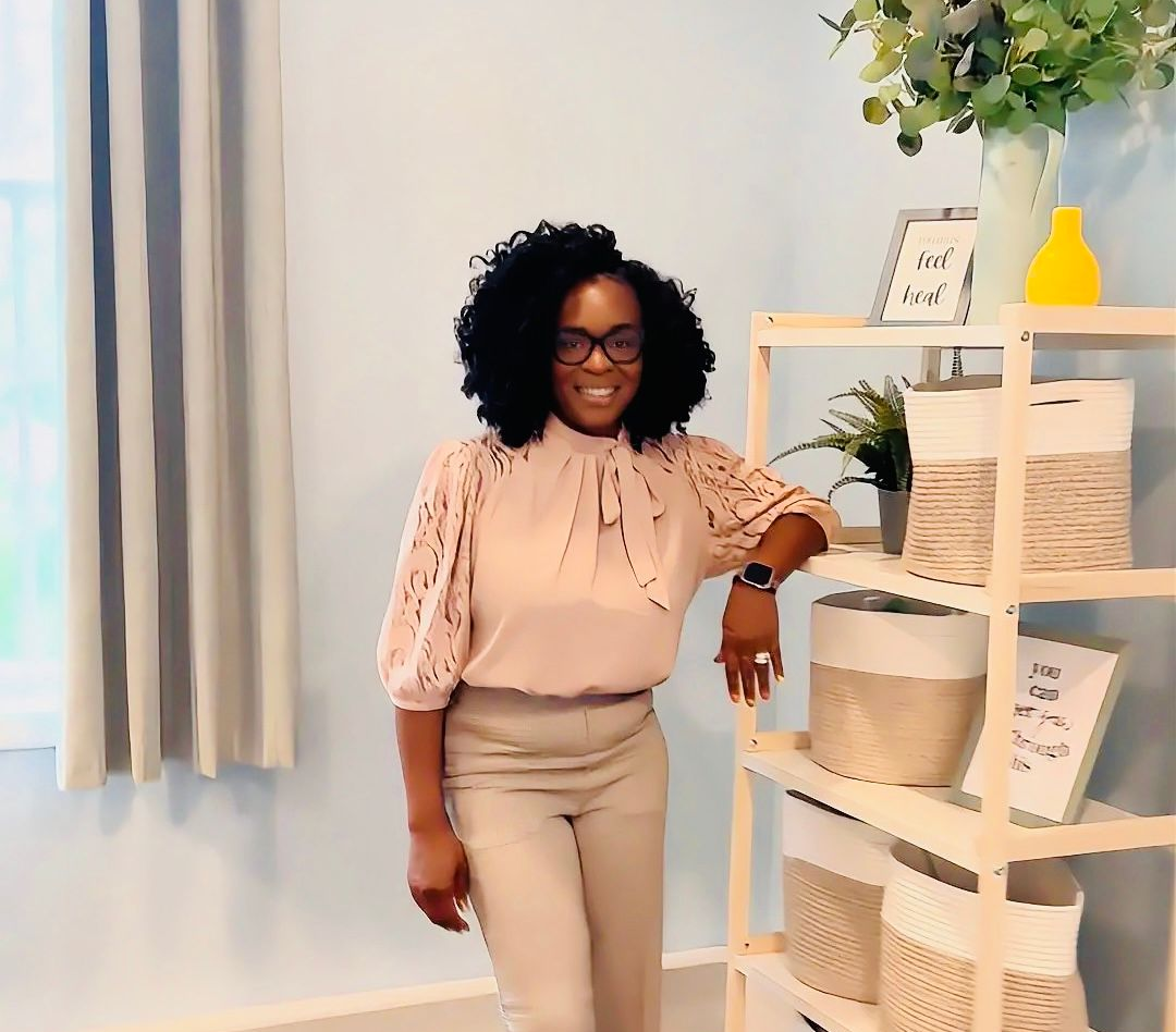 Woman posing beside white shelving and curtains in a bright room, wearing a blush blouse and beige pants.