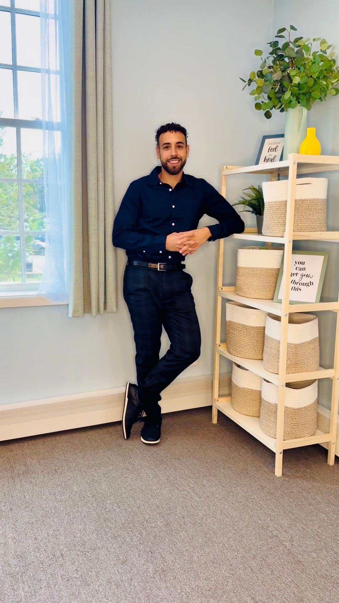Man smiling at a desk with a laptop in a bright office, wearing a dark outfit.
