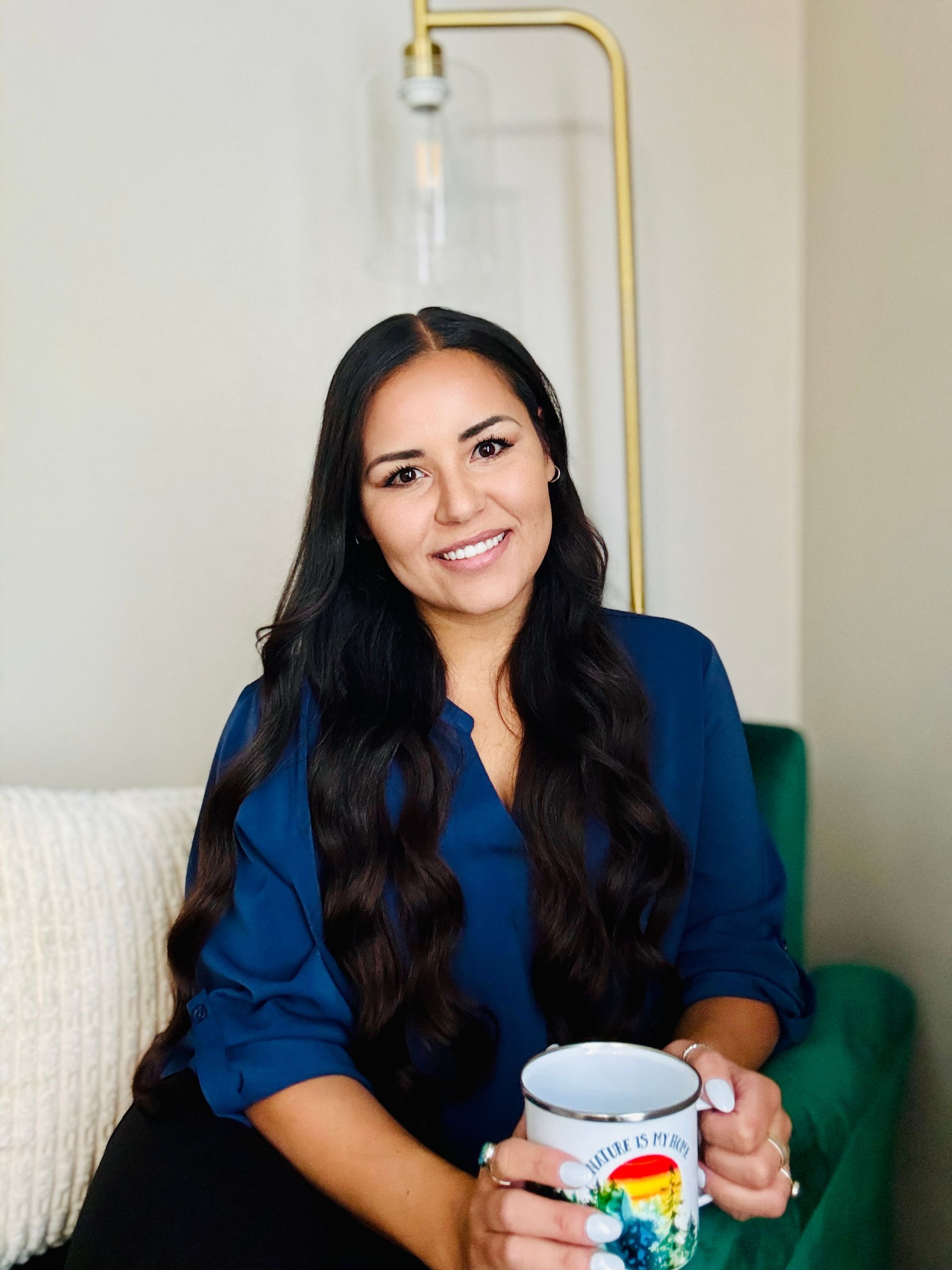 Woman smiling on a couch, holding a colorful mug in a bright room with an IV stand behind her.
