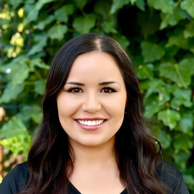 Smiling woman with dark hair in a black top, standing before a leafy green background
