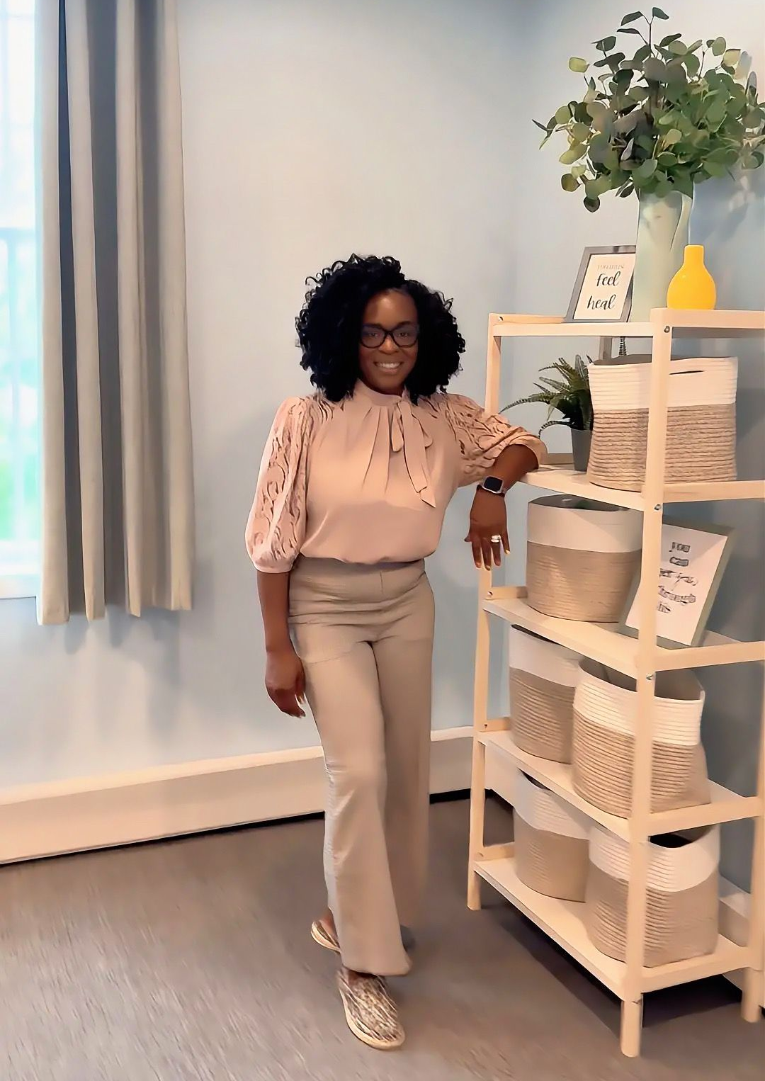 Woman standing beside a white shelving unit in a bright room with curtains and a plant.