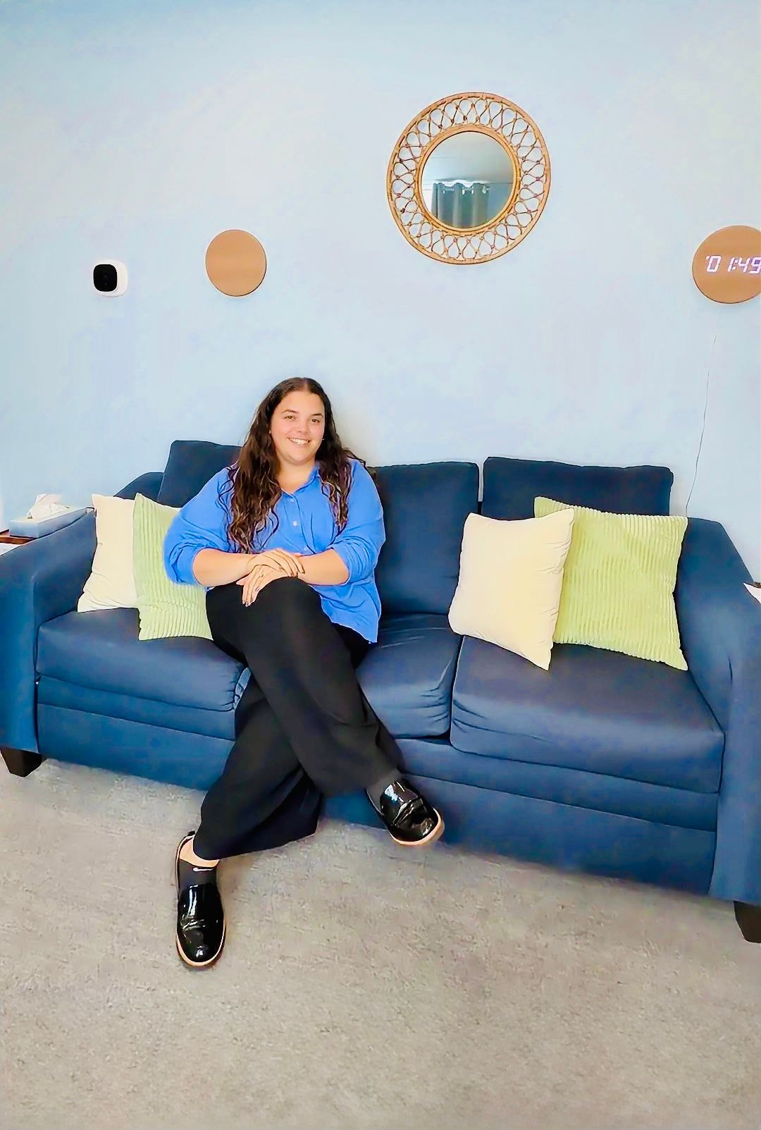 Woman sitting on a blue couch in a light blue room, smiling with crossed legs.
