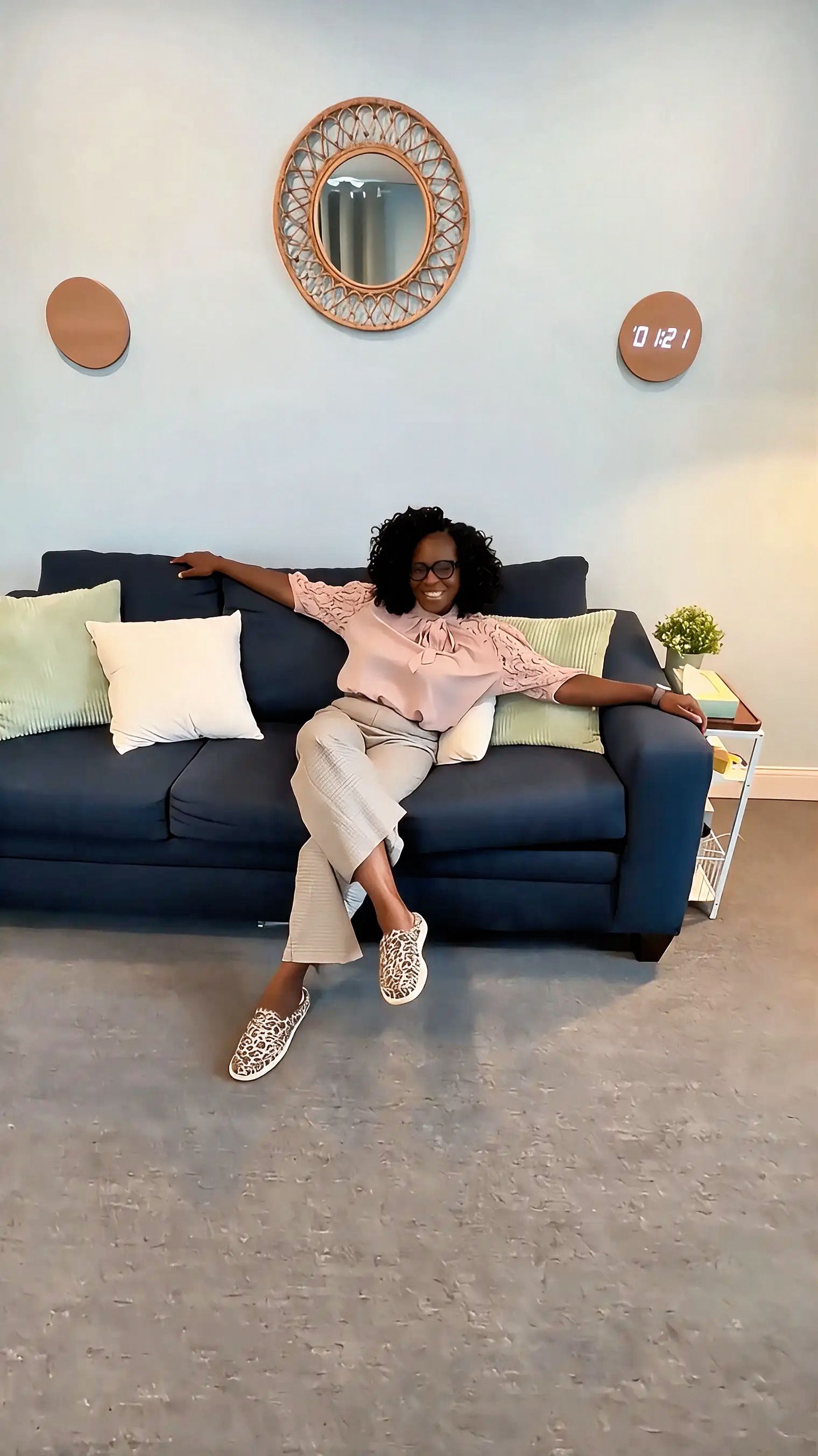 Person lounging on a blue couch in a bright living room, arms outstretched with pillows and wall decor behind.