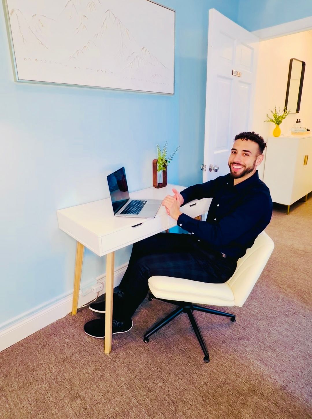 Person sitting at a desk in a bright home office, smiling beside a laptop and a whiteboard.