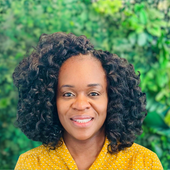 Smiling woman with curly hair in a yellow patterned blouse against a green leafy background