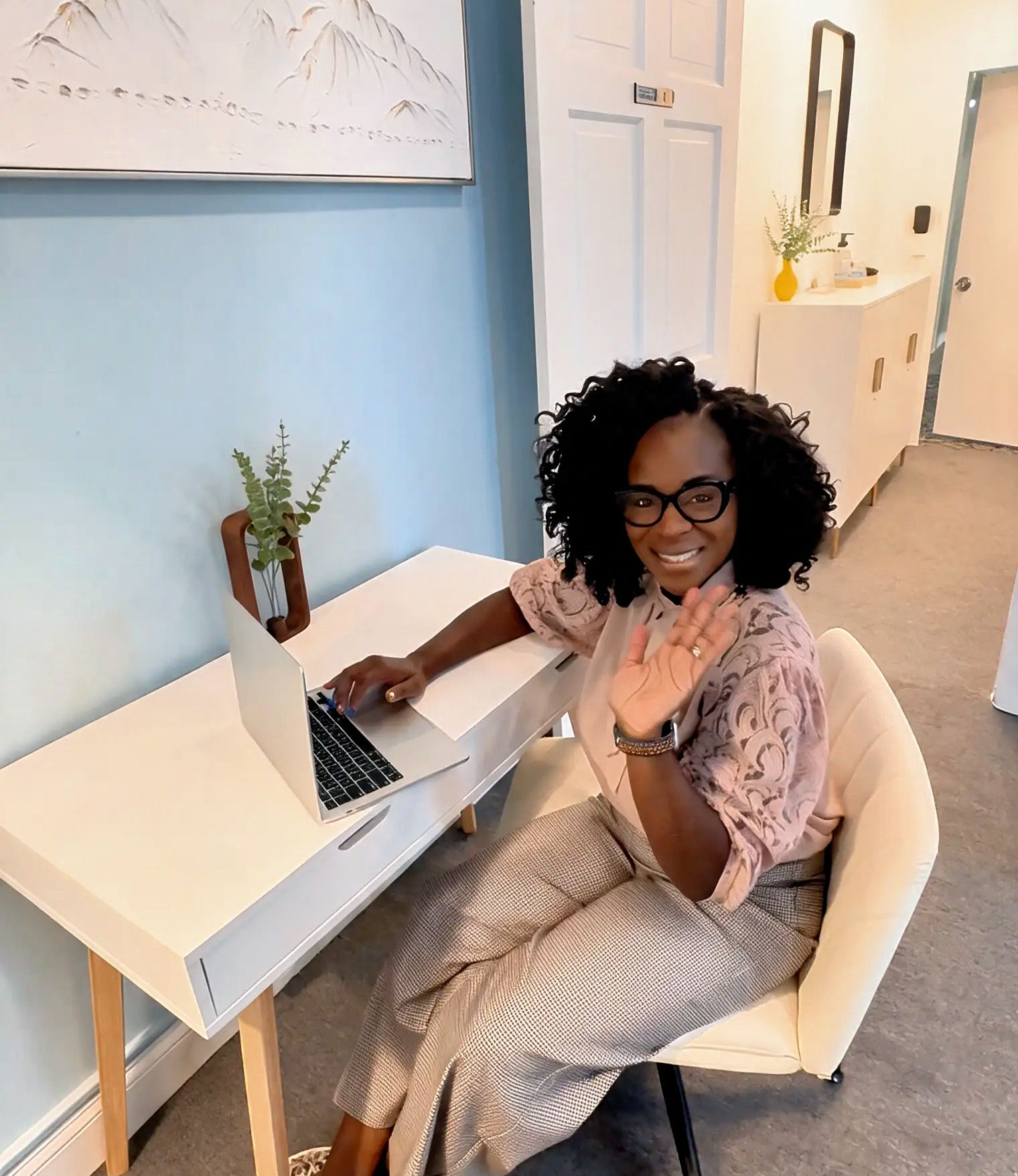 Woman smiling at a desk with laptop in a bright office, waving at the camera.