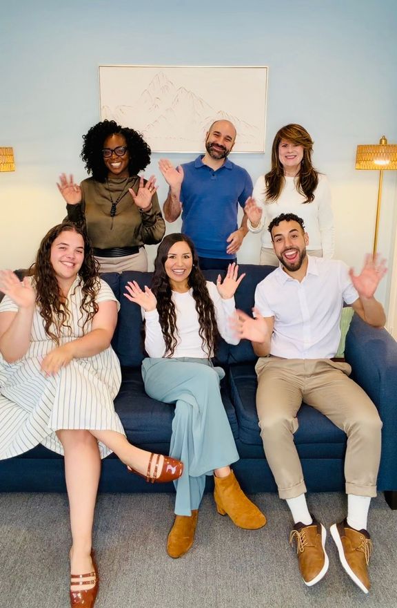 Six smiling people wave from a living room couch and chair set.
