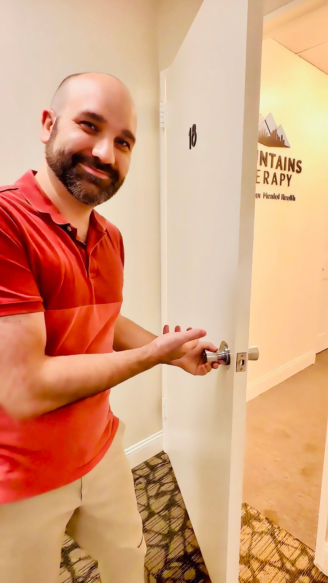 Smiling man in an orange polo opens a white door with a logo on it