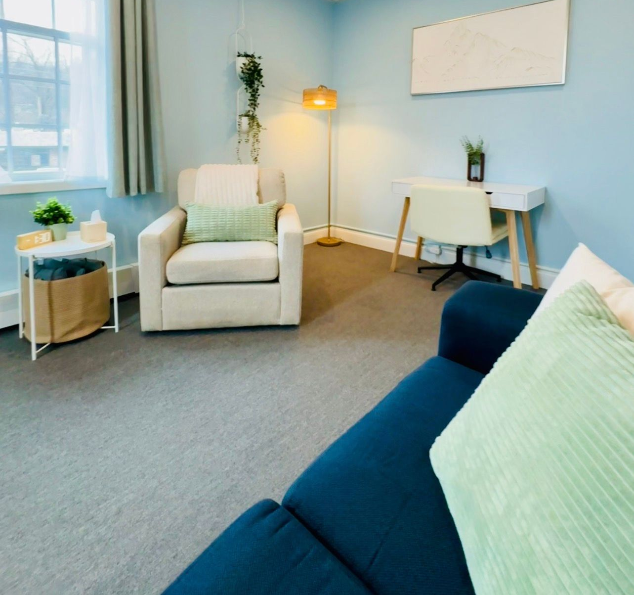 Cozy living room with white armchair, blue sofa, floor lamp, desk, and soft green accents.
