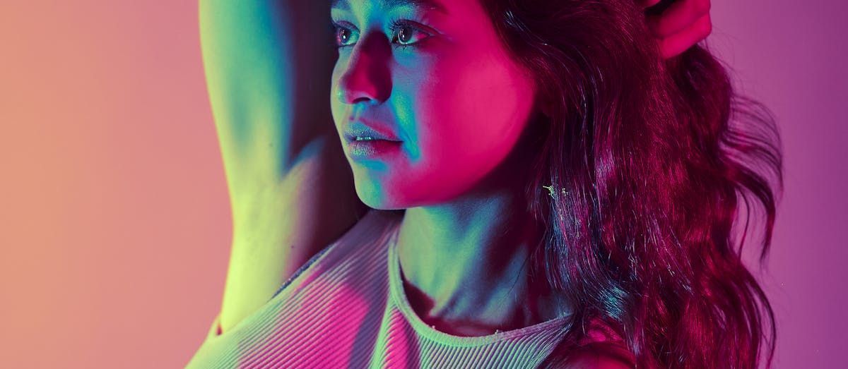 A woman with colorful lighting on her face and hair, arm raised up, looking away.