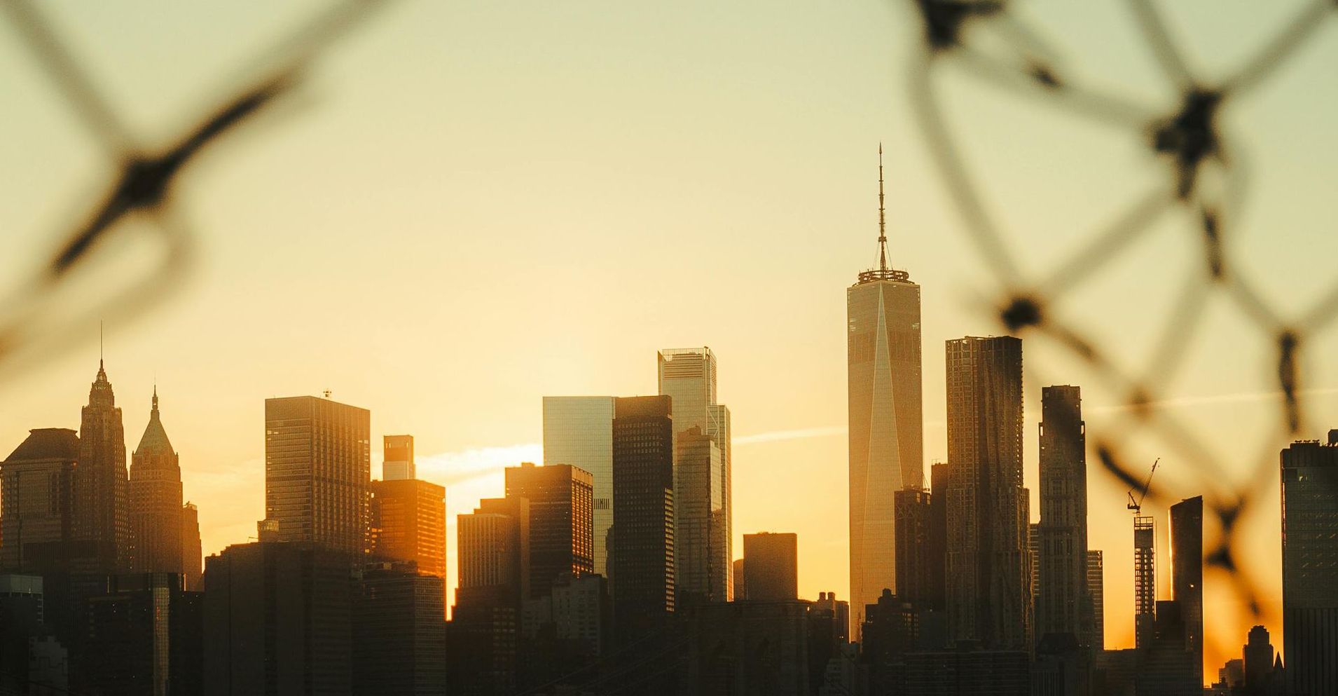New York City skyline at sunset, viewed through a blurry chain-link fence.