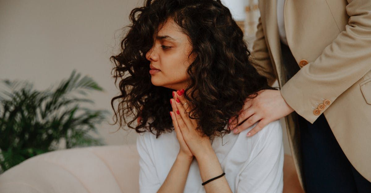 Woman with curly hair, eyes closed, praying hands, comforted by someone with a hand on her shoulder.