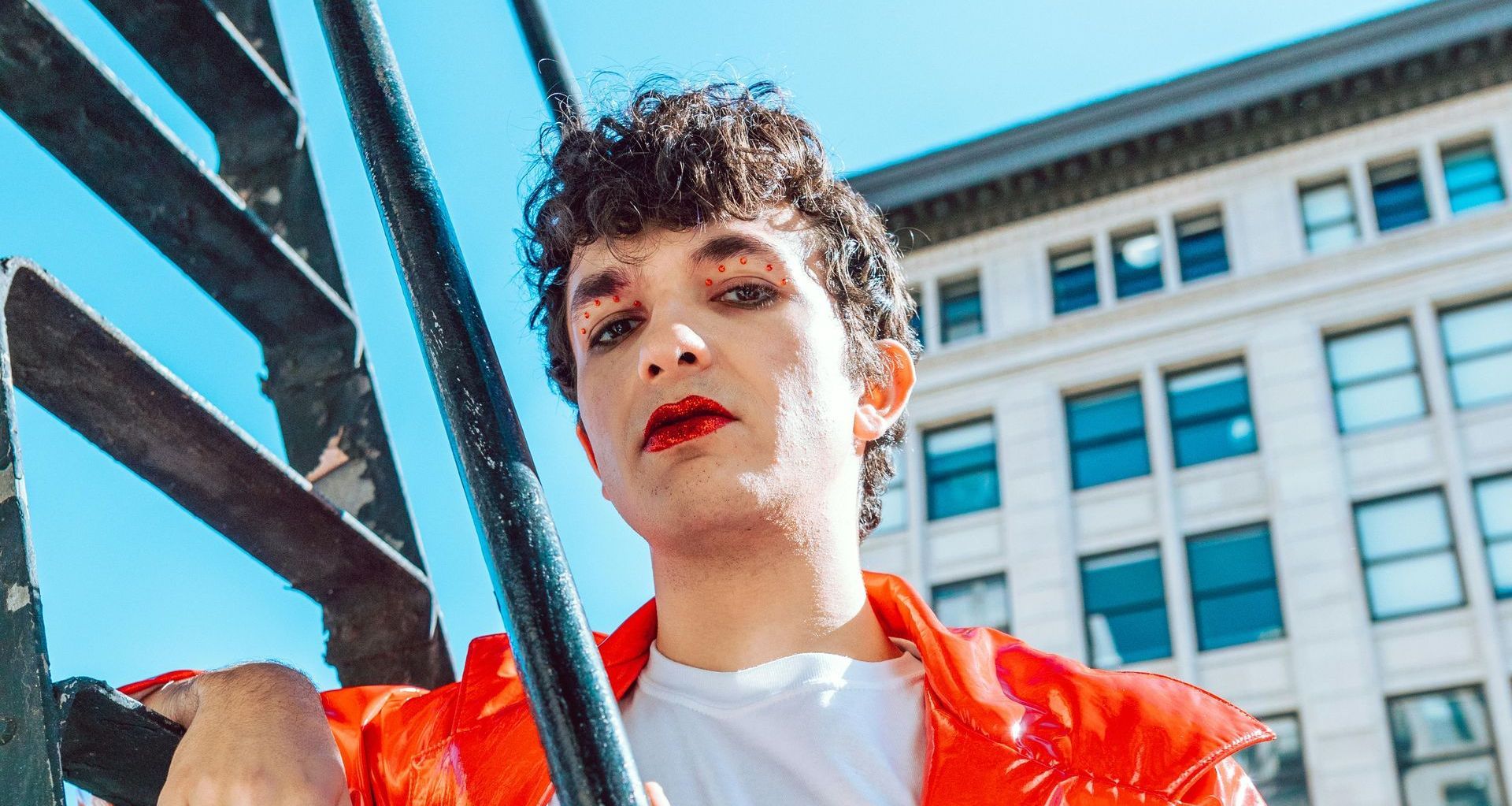 A person with makeup in a red jacket leans against a dark railing outdoors with a building in the background.