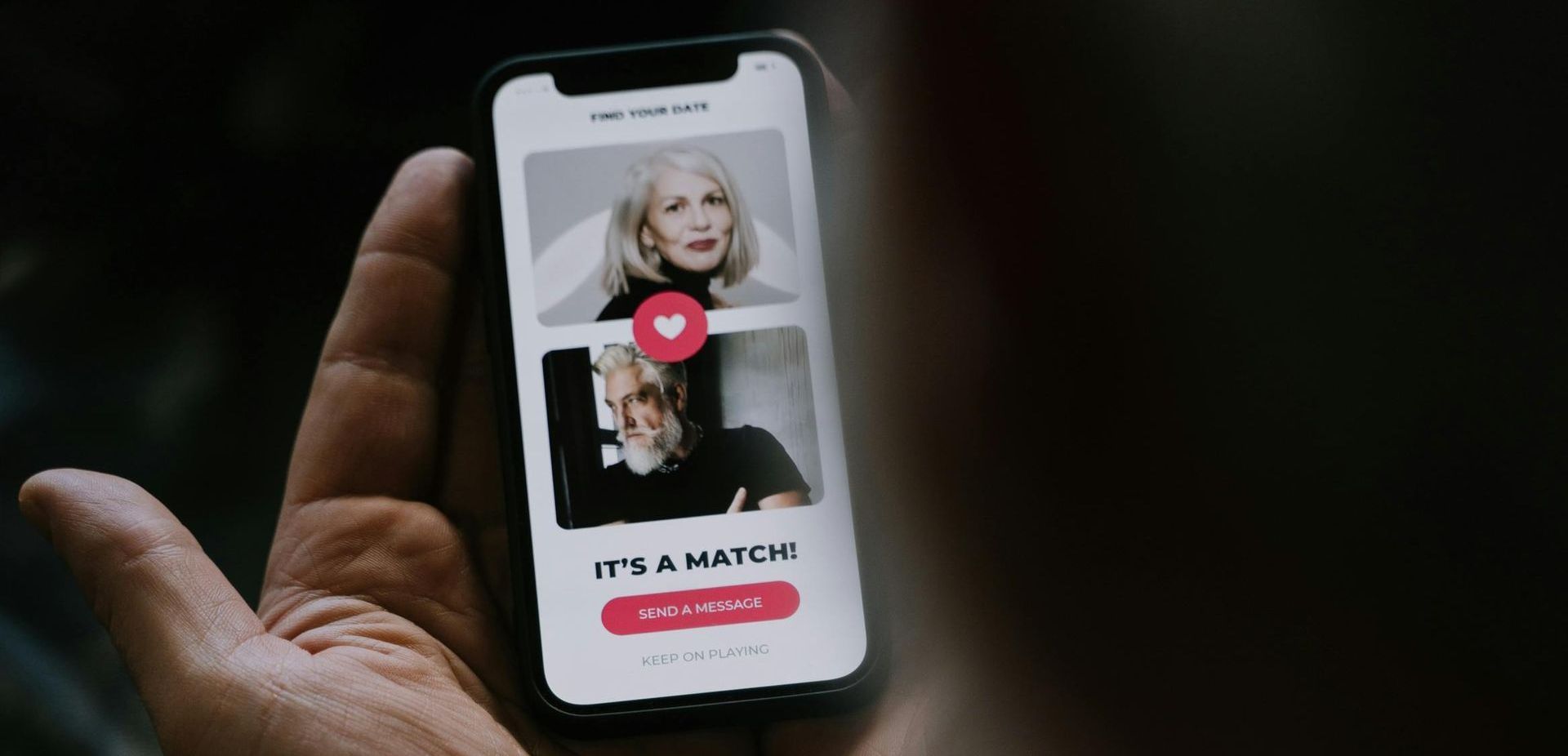 A hand holds a phone displaying a dating app. Two older women are shown with a heart indicating a match.