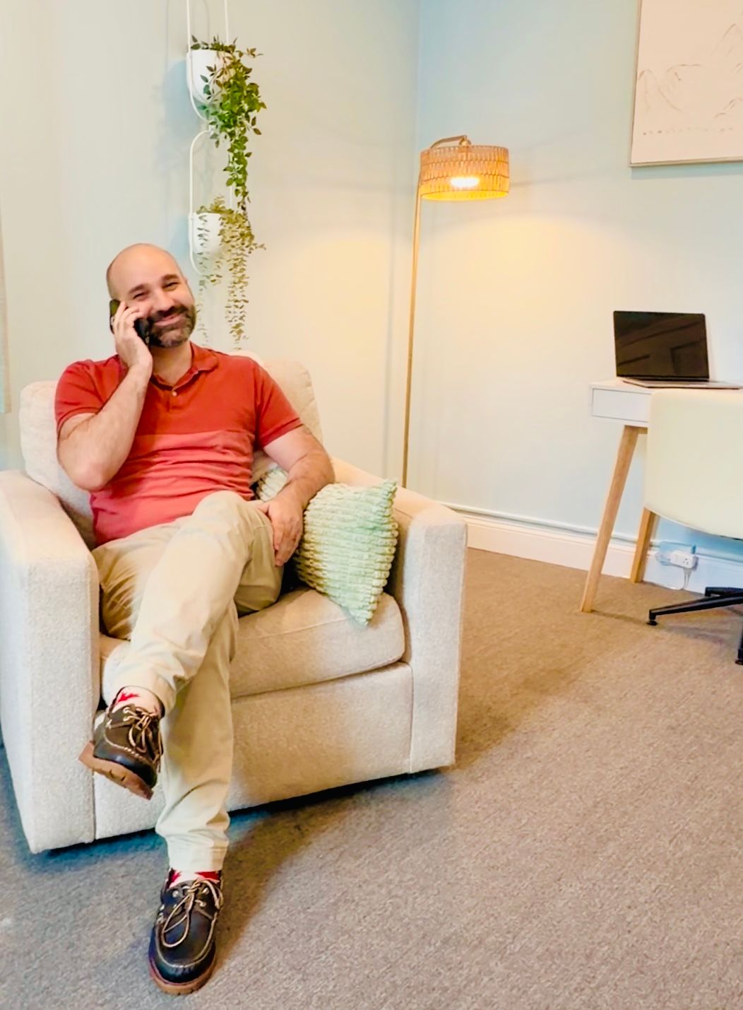 Man in a coral shirt sitting in a beige armchair, smiling on a phone in a bright living room.
