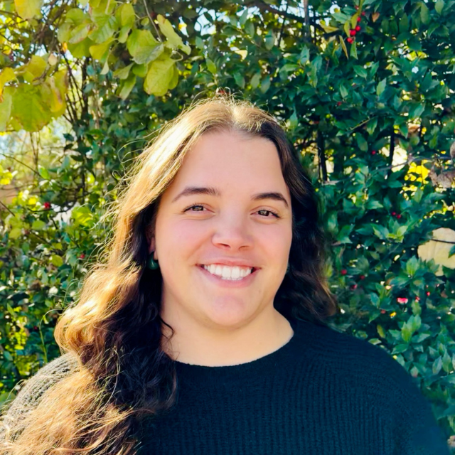 Smiling person with long wavy brown hair wearing a black sweater, standing in front of leafy green trees.