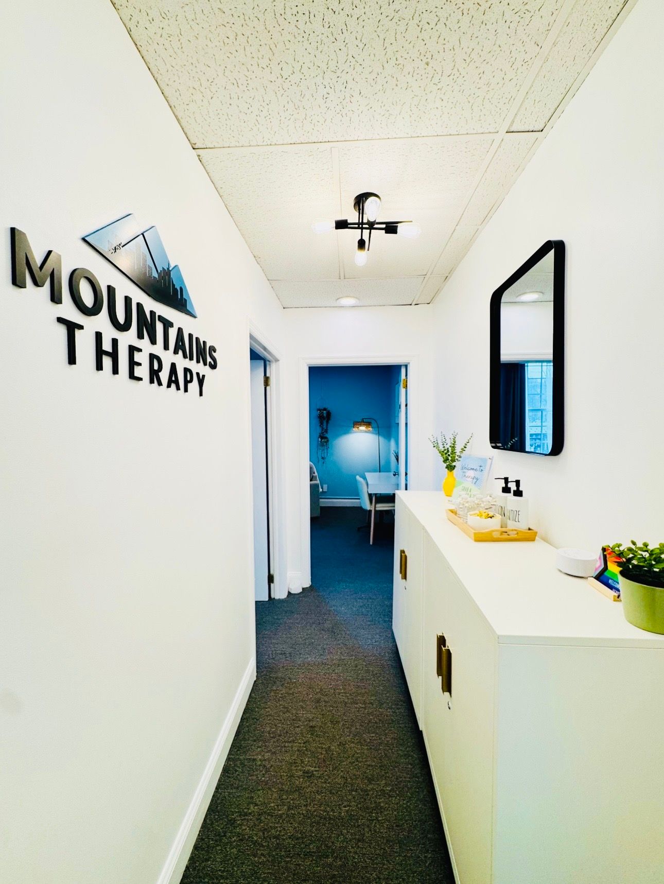 Hallway leading to therapy room; white walls, cabinet, mirror, Mountains Therapy sign, gray carpet.
