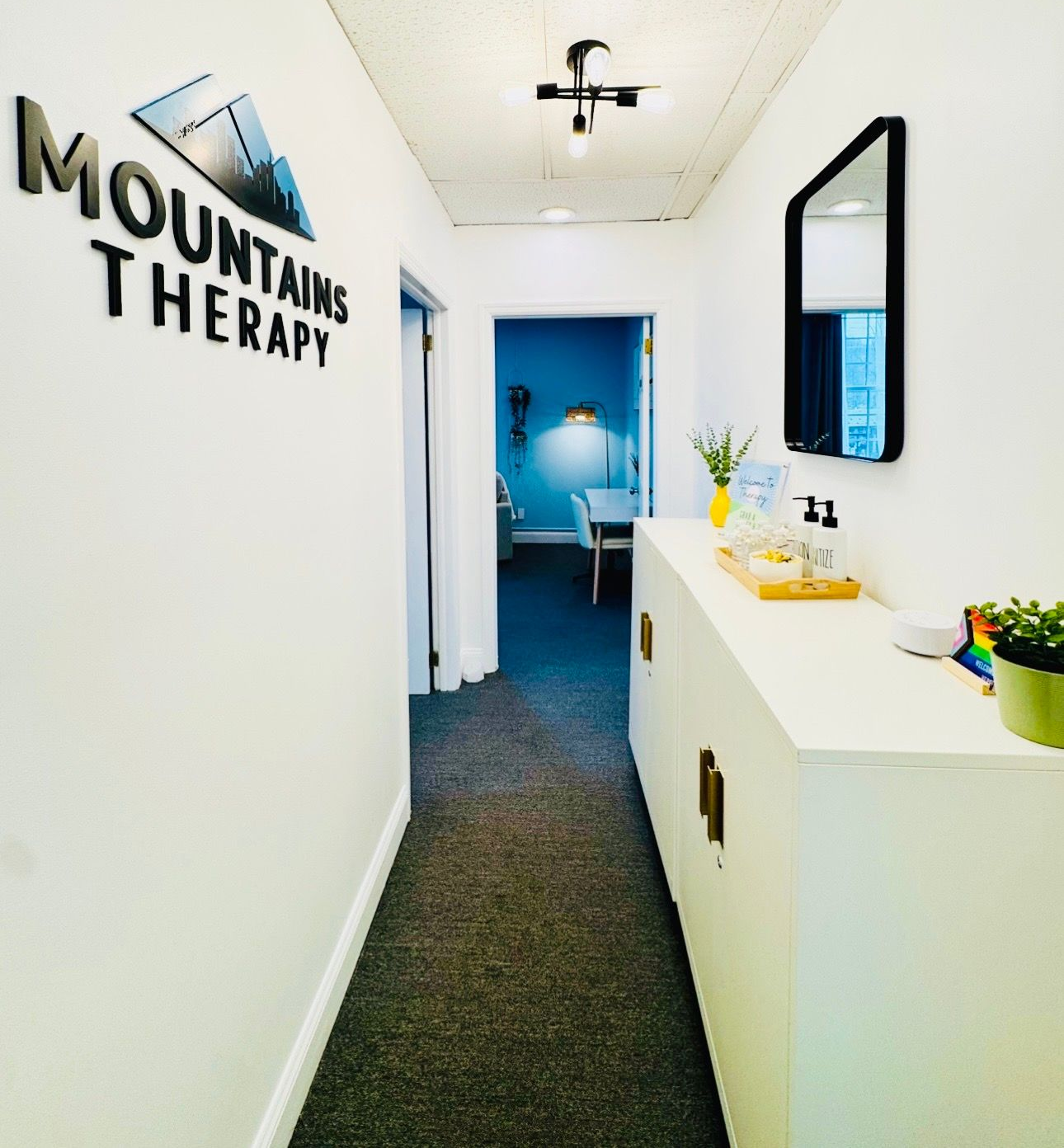 Narrow white hallway leading to a blue room, with “Mountain Therapy” wall sign and a small reception counter.