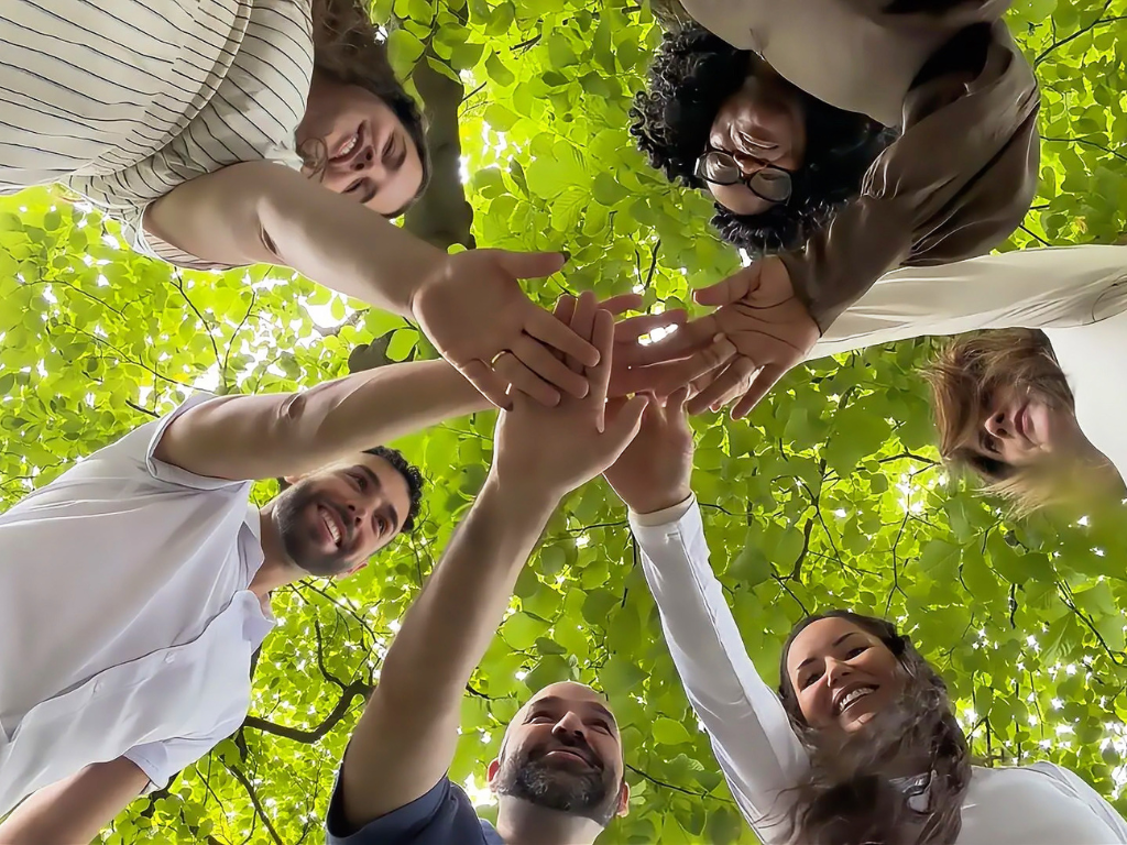 Six people stand in a circle, stacking hands together against a leafy green sky.