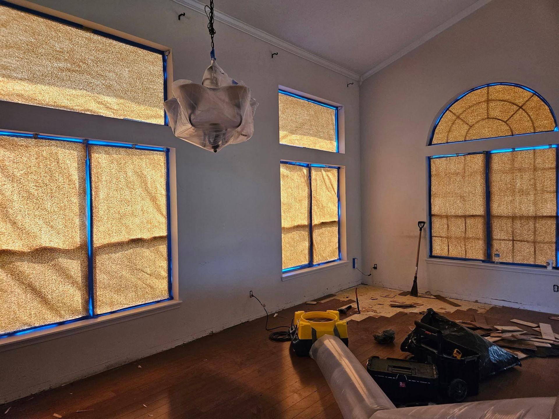 Partially renovated room with covered windows, a hanging light, and tools and debris on the floor.