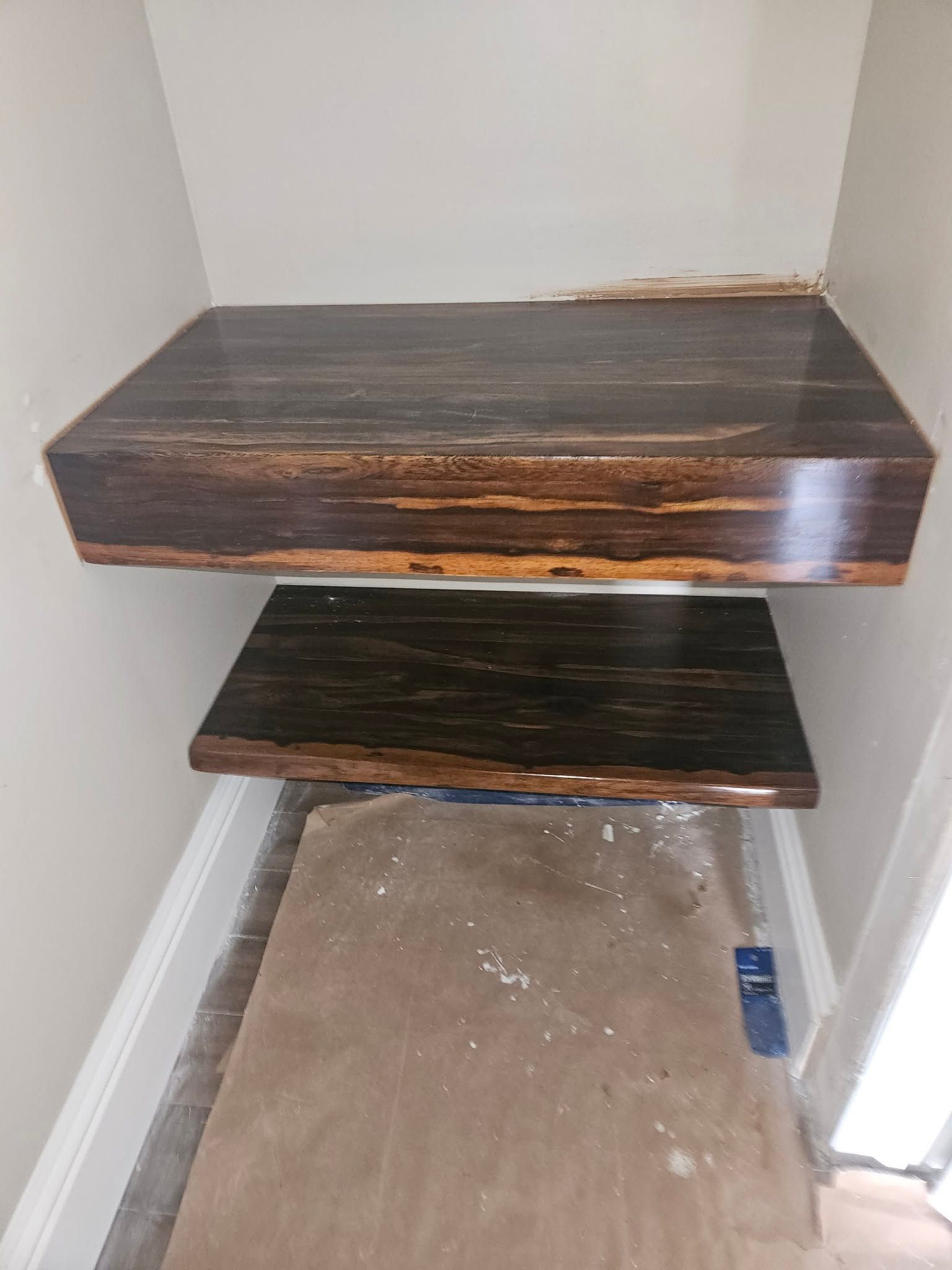 Two dark wooden floating shelves mounted in a small corner with a protective floor covering below.