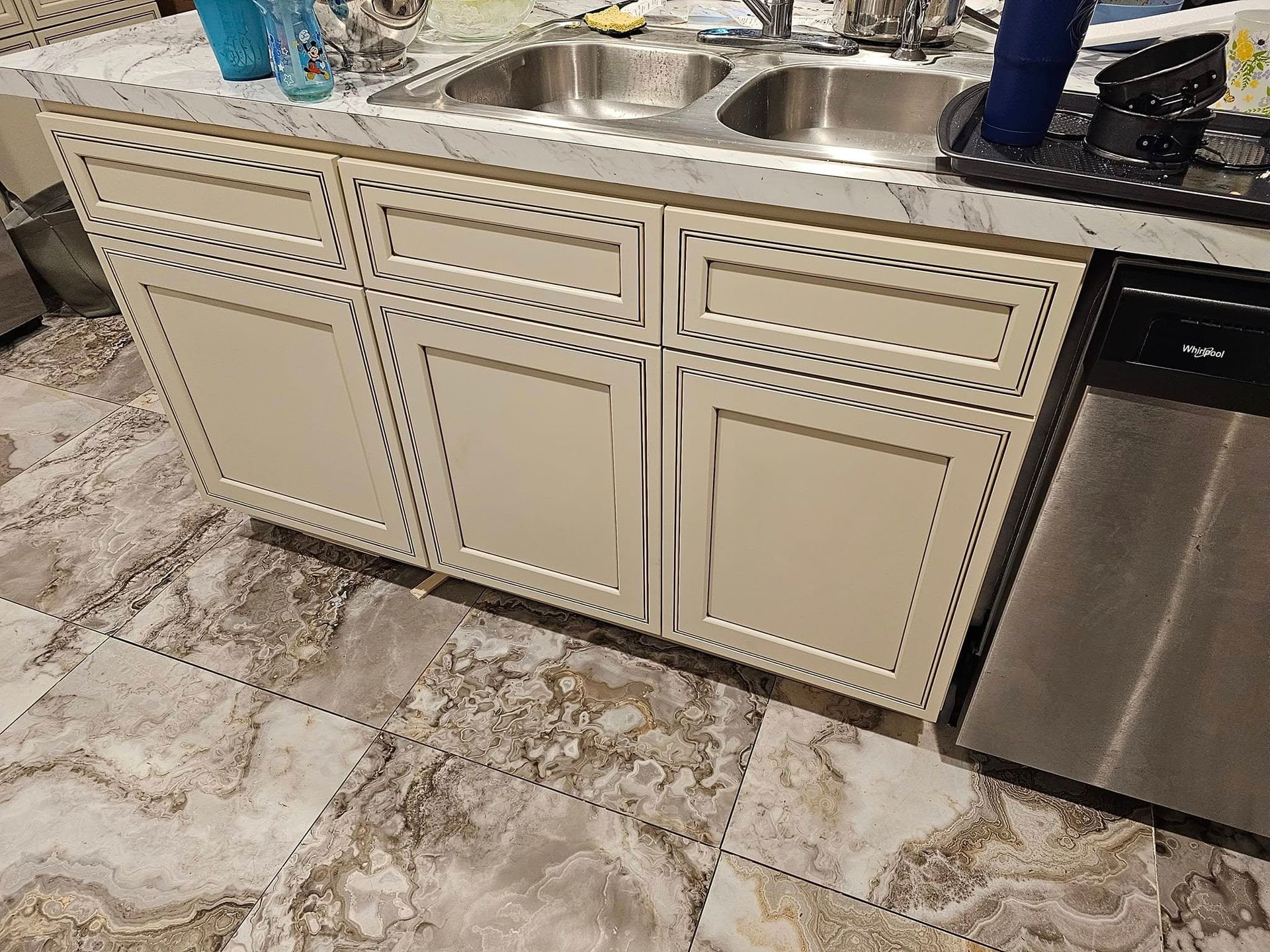 Kitchen sink and cream cabinets with decorative trim, beside a stainless steel dishwasher and patterned tile floor.
