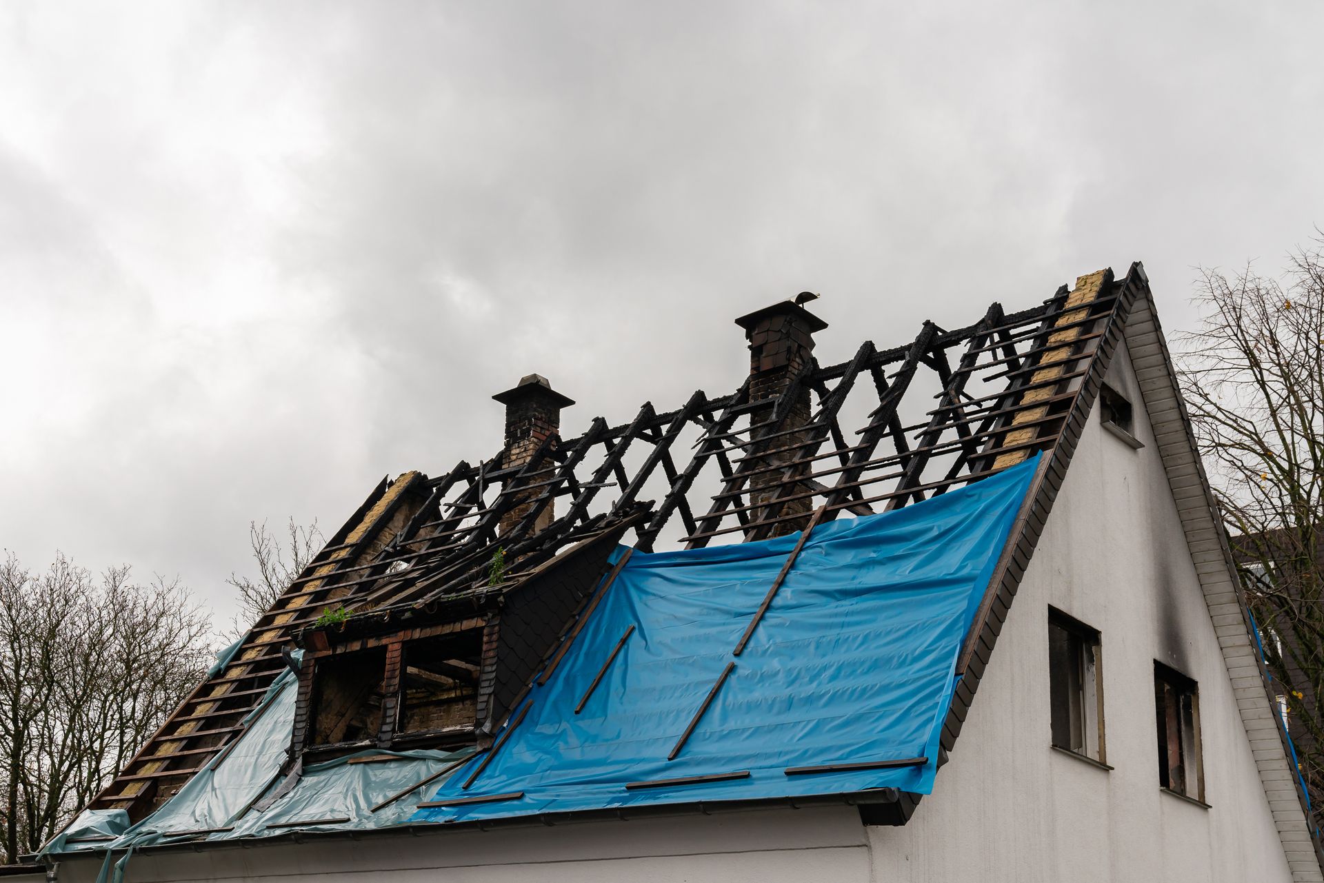 Fire-damaged house roof with exposed rafters and blue tarp covering part of the roof.