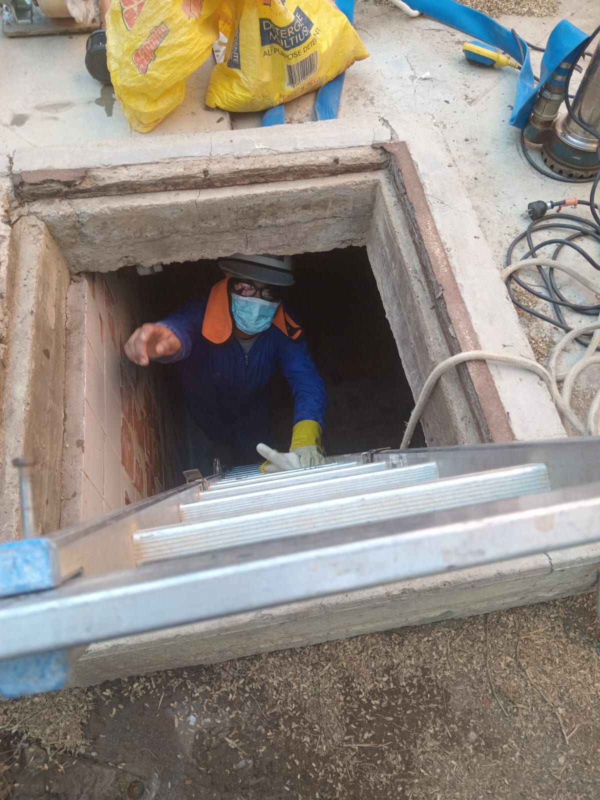 Un trabajador sale de un estrecho pozo subterráneo, con mascarilla y guantes, con una escalera en primer plano.