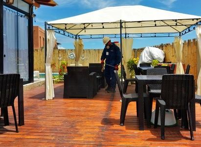 Terraza de madera al aire libre con mesas y sillas negras, y un trabajador limpiando bajo un toldo blanco.