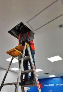 Un trabajador, subido a una escalera, repara un panel del techo de una oficina con luces empotradas.