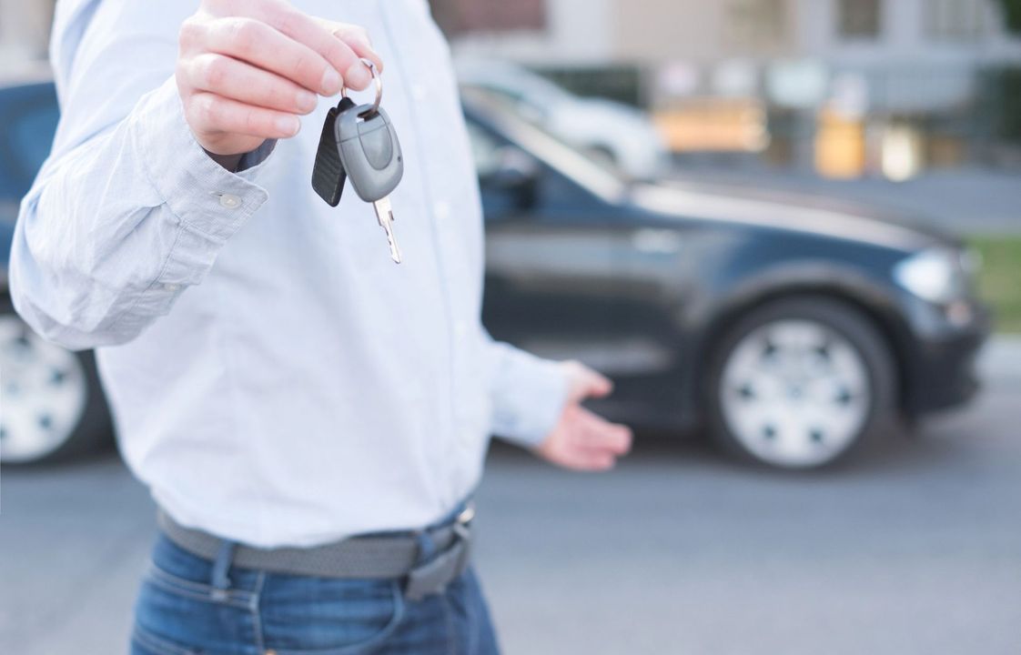 Una persona tiene in mano le chiavi di un'auto davanti a un'auto nera parcheggiata.