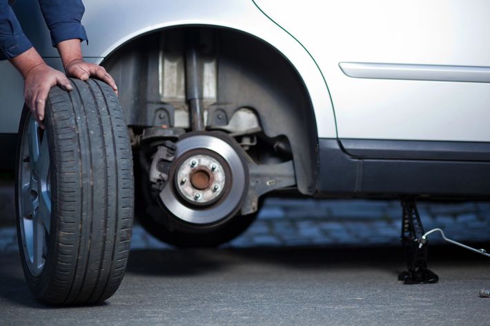 L'auto viene sollevata con il cric e la ruota anteriore rimossa; il meccanico tiene una ruota di scorta accanto al gruppo freno esposto.